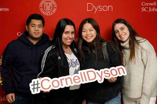 Four people smile in front of a Dyson step and repeat holding a “#CornellDyson” sign.