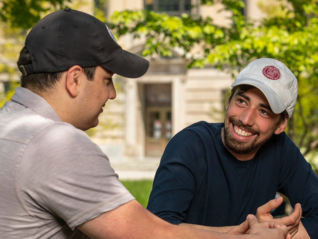 Two men in baseball hats sit outside talking.